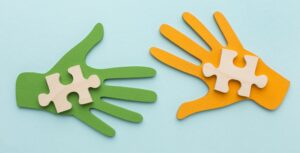 Symbolbild - eine grüne und eine gelbe Hand aus Holz mit einem Puzzelteil geschmückt die sich die Hand reichen